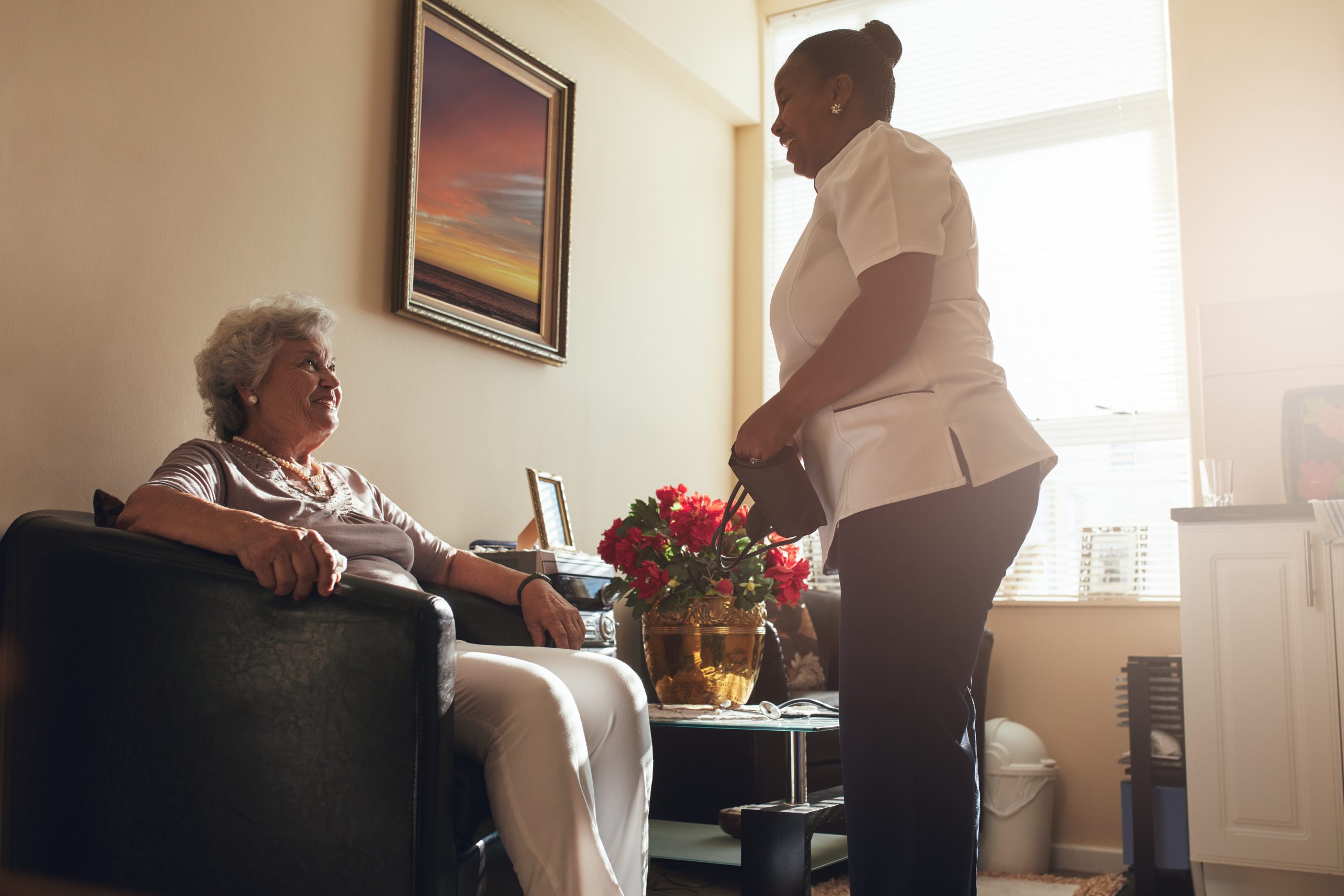 Senior woman sitting on a chair at home with female caregiver standing by. Female nurse visiting senior patient for checking blood pressure.